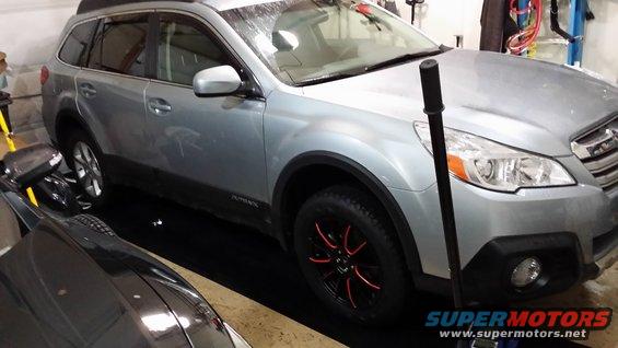 Silver 2014 Subaru Outback parked indoors with black wheels and red accents. Another car is partially visible in the foreground