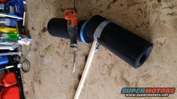 Reciprocating saw cutting a rolled black mat secured with tape and a wooden brace