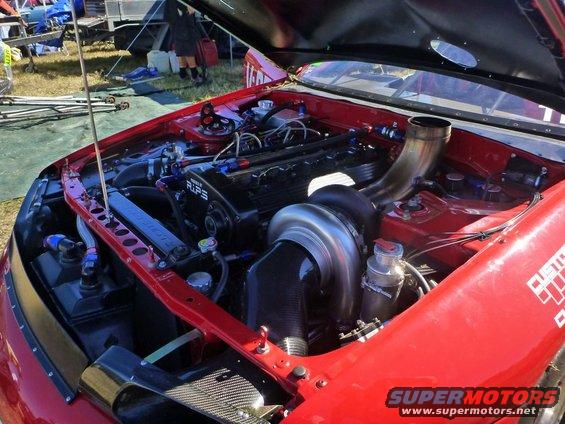 Engine bay of a red car with a large turbocharger and custom piping. Text on the fender reads 'CUSTOM'