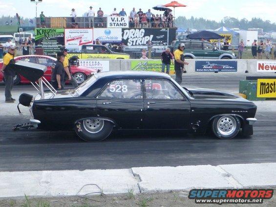 Black drag car with number 528 on the window, at a drag strip with spectators in the background
