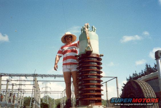 Man in striped shirt and hat stands by large electrical insulator at substation. Tire visible on right
