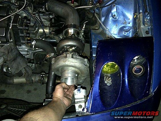 Blue car engine bay with turbocharger, hoses, and a hand adjusting a part. Yellow fog light visible