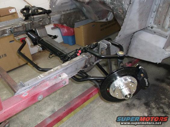 Car chassis showing front suspension, brake caliper, and rotor assembly. Some body panels are visible in the background