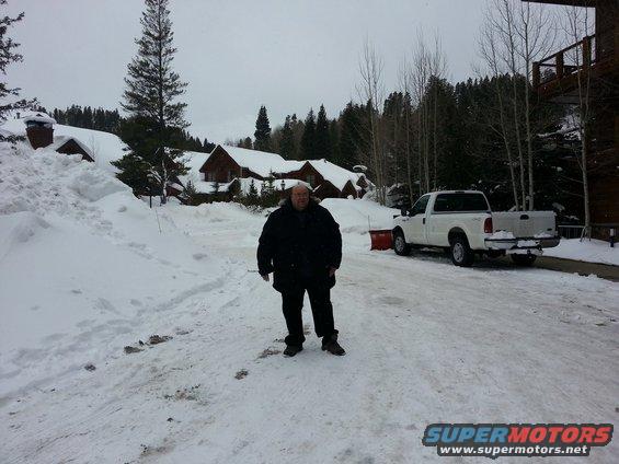 White pickup truck with a red snow plow attachment parked in a snowy area with buildings and trees