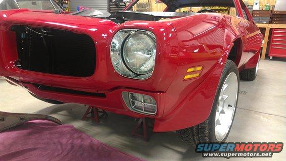 Front end of a red muscle car on jack stands, hood up, showing the grille and headlights