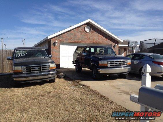 Two Ford trucks parked in front of a garage, one is a dark color with '1075' written on the windshield, the other is blue. A silver sedan is parked to the right
