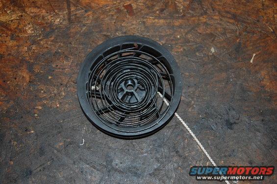 Black recoil starter assembly with a coiled spring and pull rope, on a wooden surface