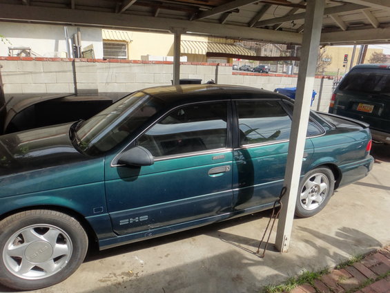 1993 Ford Taurus sedan, dark green, parked under a carport. Side view shows the front and rear wheels, doors, and windows