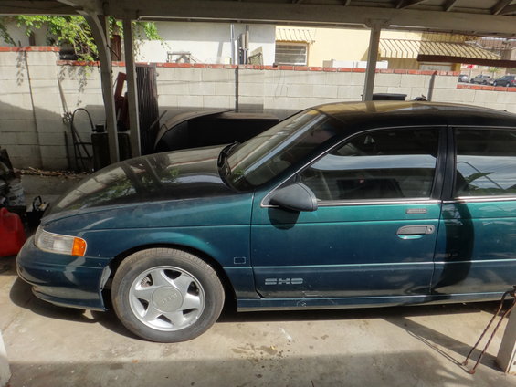 Side view of a 1993 Ford Taurus sedan parked under a carport. It has a dark green body with a black roof and silver wheels