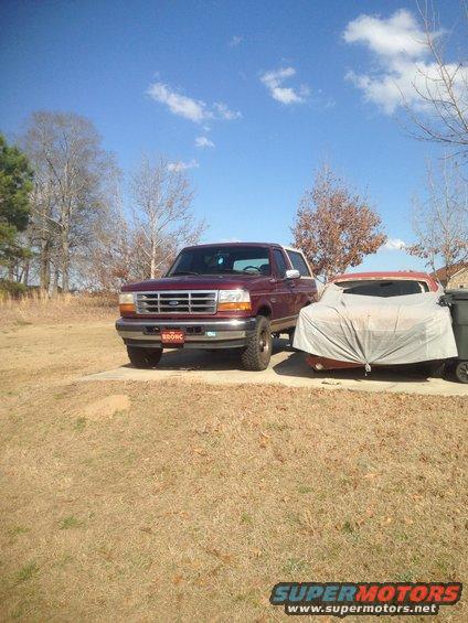 Maroon 1996 Ford Bronco parked on concrete, next to a tarp-covered car