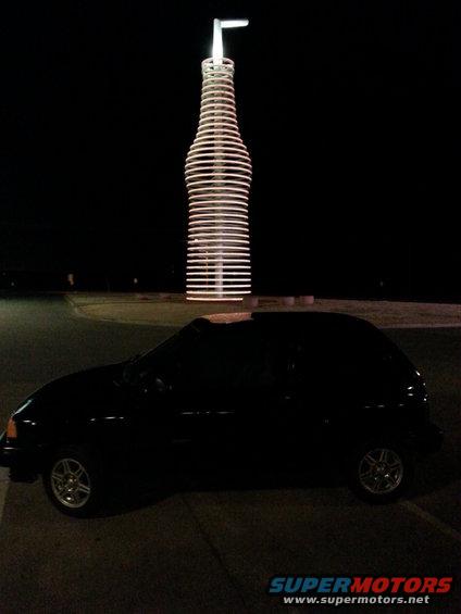 black hatchback parked at night with a large neon bottle sculpture behind it