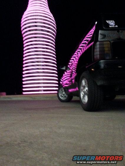 Black car with pink zebra stripe trailer parked next to a neon pink bottle sculpture