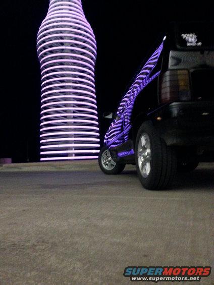 Black hatchback with purple zebra stripes parked at night next to a large illuminated bottle structure