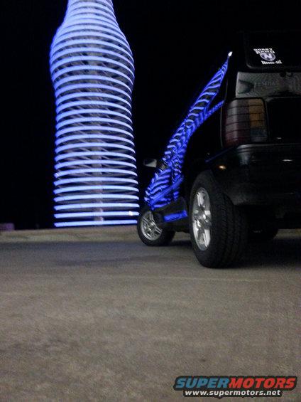 Blue LED-lit scooter parked next to a tall, illuminated bottle sculpture at night
