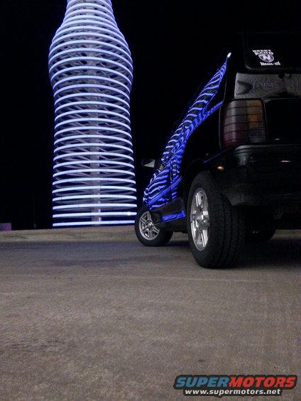 Black car with blue neon lights on its side parked near a giant illuminated bottle structure
