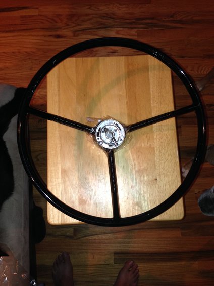Black three-spoke steering wheel with chrome center hub, resting on a wooden table
