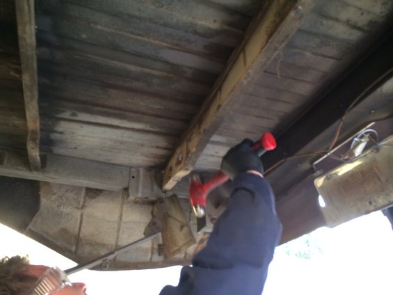 Underside of a wooden truck bed with a person in black gloves hammering a metal support