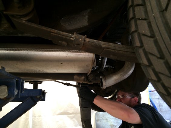 Underside of a vehicle with a mechanic working on the exhaust system, showing a leaf spring and muffler