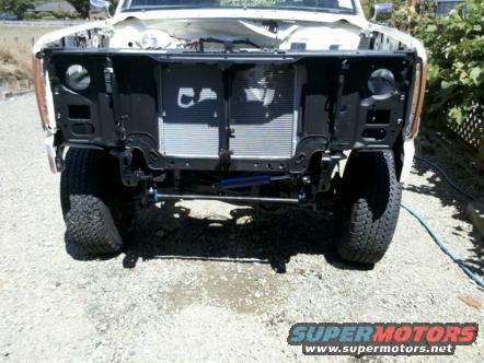 Front end of a white truck, grille removed, showing radiator and suspension components. Tires are large and off-road style