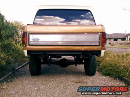 Gold and white Ford Bronco rear view. Visible chrome trim, taillights, and differential. Gravel road