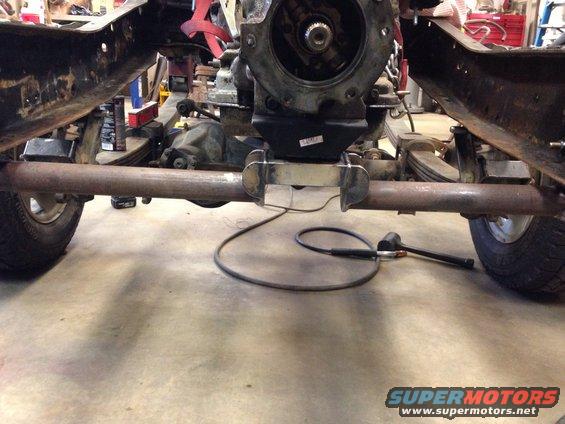 Underside view of a vehicle frame with drivetrain components, leaf springs, and exhaust pipe. Welding equipment is visible
