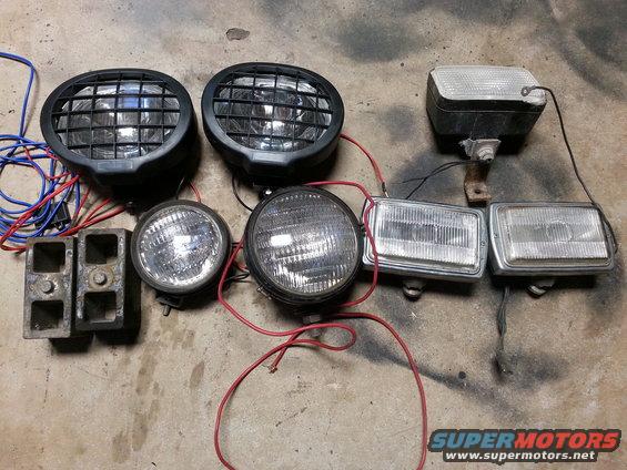 Collection of automotive lights including two large round lights with grilles, two smaller round lights, and two rectangular lights, plus two metal brackets