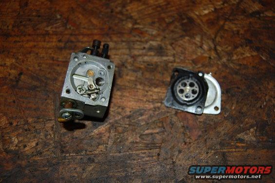 Two parts of a carburetor are on a wooden surface. One is a metal assembly with levers and screws. The other is a black disc with holes