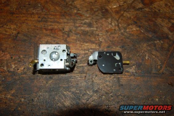 Two carburetor components on a wooden surface. One is a silver metal assembly with brass fittings. The other is a black plate with metal accents