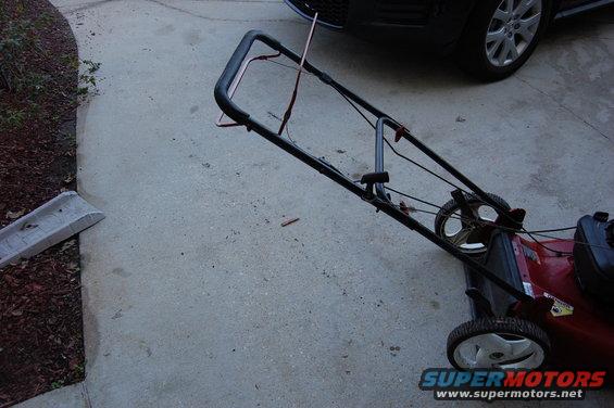 Red push mower with black handle and wheels on concrete. Part of a dark vehicle is visible in the background