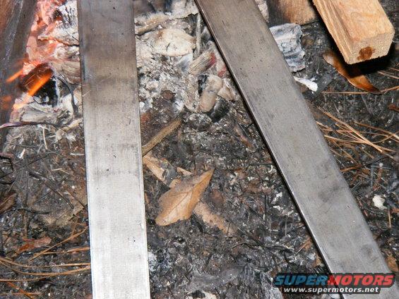 Two metal bars lie across campfire ashes with burning embers visible on the left