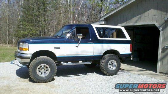 Blue and white 1994 Ford Bronco with lifted suspension parked next to a garage