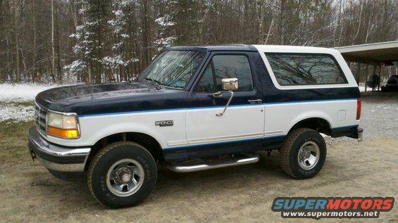 Two-tone blue and white 1994 Ford Bronco parked on dirt with trees in background