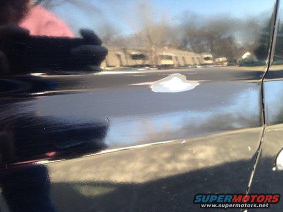 Close-up of a dark vehicle's side panel showing rust spots and primer. Reflection of buildings in the background