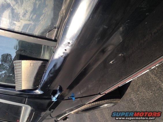 Close-up of a black vehicle's side panel showing drilled holes and some blue tape
