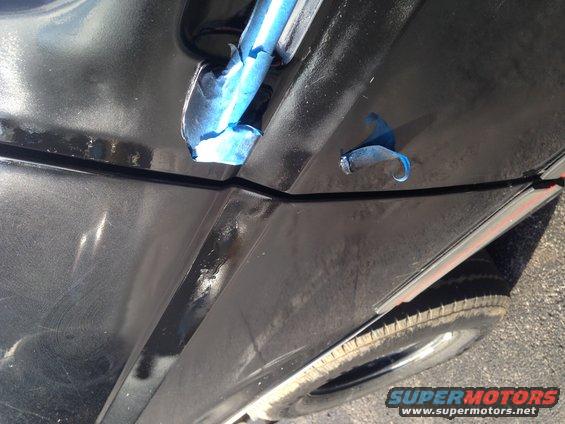Close-up of a black car's door seam with blue painter's tape. A tire is visible at the bottom