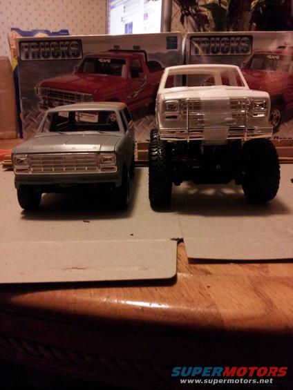Two model trucks, one gray and one white, sit on a table with model truck boxes in the background