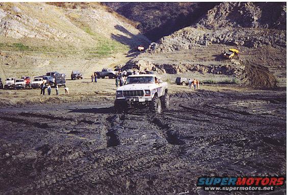 White off-road truck with large tires driving through mud, front view. Grille indicates 1980s model