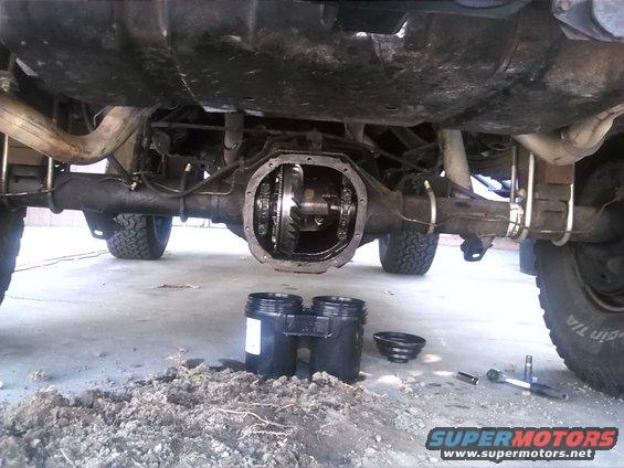 Underside of a vehicle showing the rear differential with the cover removed. Tools and containers are on the ground
