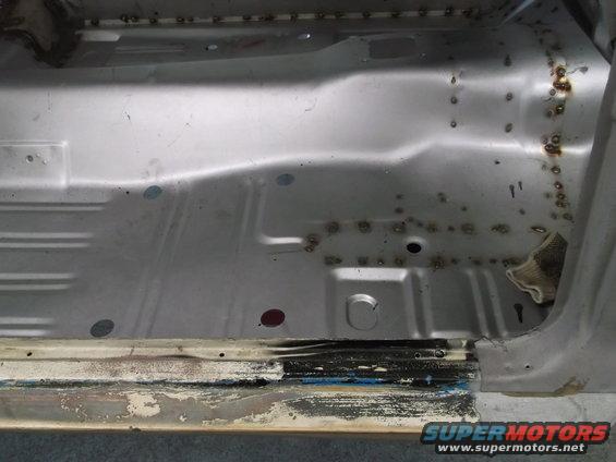 Car floor pan with visible rust and repair welds. The metal is bare and shows signs of previous bodywork