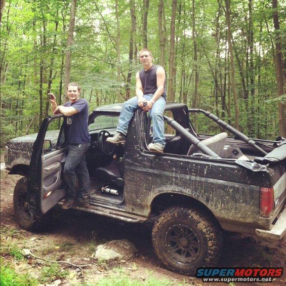 Black Ford Bronco with two men, one standing by the open driver door and another sitting on the roof, in a wooded area
