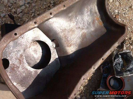 Rusty metal oil pan with circular cutout and bolt holes, next to engine parts