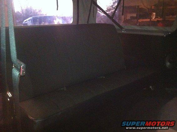 Dark rear bench seat with seatbelts, viewed from inside a vehicle. A second vehicle is visible through the rear window