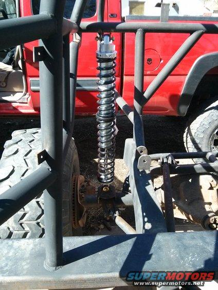 Close-up of a heavy-duty shock absorber and suspension components on a custom off-road vehicle, with a red truck visible in the background
