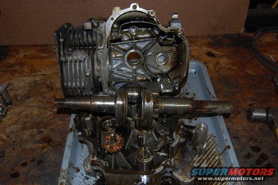 Engine crankshaft and block disassembled in a gray tray, with oil and debris. A hammer is visible to the right