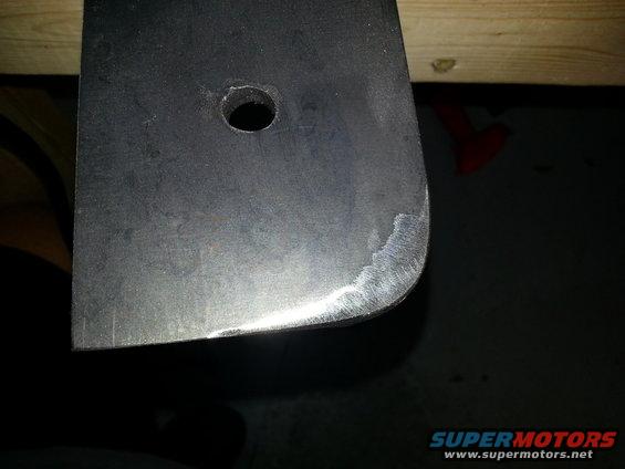 Close-up of a dark metal bracket with a hole and rounded corner. Text at bottom says SUPERMOTORS