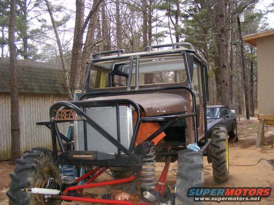 Custom off-road vehicle with large tractor tires, roll cage, and exposed engine bay. A dark sedan is visible behind it