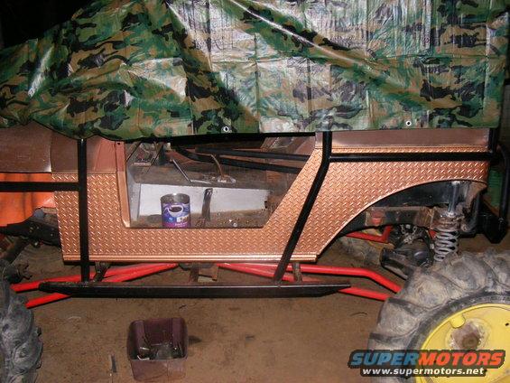 Custom off-road vehicle with diamond plate accents and a camouflage tarp covering the top. Red roll cage and suspension components are visible