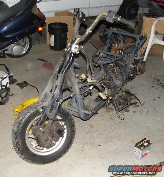 Scooter frame, engine, and wiring laid out on garage floor. Another scooter is parked to the left