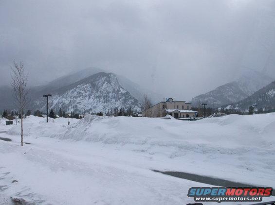 Snow-covered ground and mountains under a cloudy sky, with a building in the mid-ground