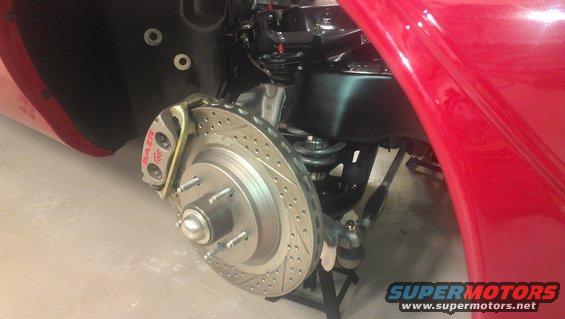 Close-up of a car's front brake rotor, caliper, and suspension components. The caliper has 'BAER' written on it